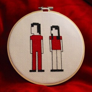 May include: A white embroidery hoop with a cross-stitch design of two pixelated figures. The figures are red with black outlines and white faces. The figures are standing side-by-side.