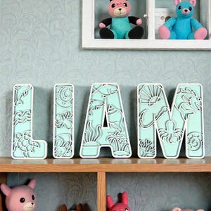 May include: A decorative name sign spelling "LIAM" in seafoam green, layered letters with white edges, featuring ocean-themed cutouts. The sign is on a wooden shelf, with pink and blue teddy bears.