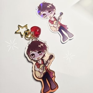 May include: A clear acrylic keychain featuring a cartoon character playing a guitar, with a gold star charm and a red cherry charm. A second sticker of the same character is also visible. The character is wearing a red vest and a white shirt.