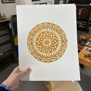 May include: A print on white paper featuring a detailed, circular design in a warm gold color. The design includes intricate patterns of lines, curves, and star-like shapes, resembling a stained-glass window or mandala. The print is held by a person.