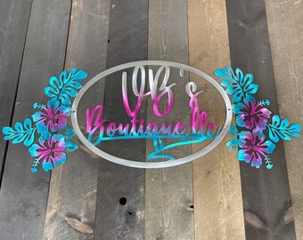 May include: A metal sign with a white oval background and the text "VB's Boutique, LLC" in pink and blue lettering. The oval is surrounded by two clusters of pink and blue hibiscus flowers.