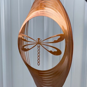 May include: A copper-colored metal wind spinner with a dragonfly cutout design. The spinner is made of multiple layers of metal that are stacked and curved to create a 3D effect.