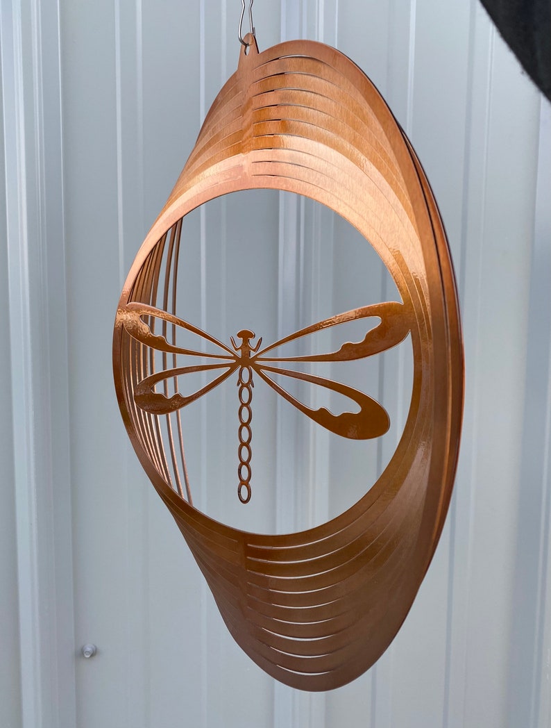 May include: A copper-colored metal wind spinner with a dragonfly cutout design. The spinner is made of multiple layers of metal that are stacked and curved to create a 3D effect.