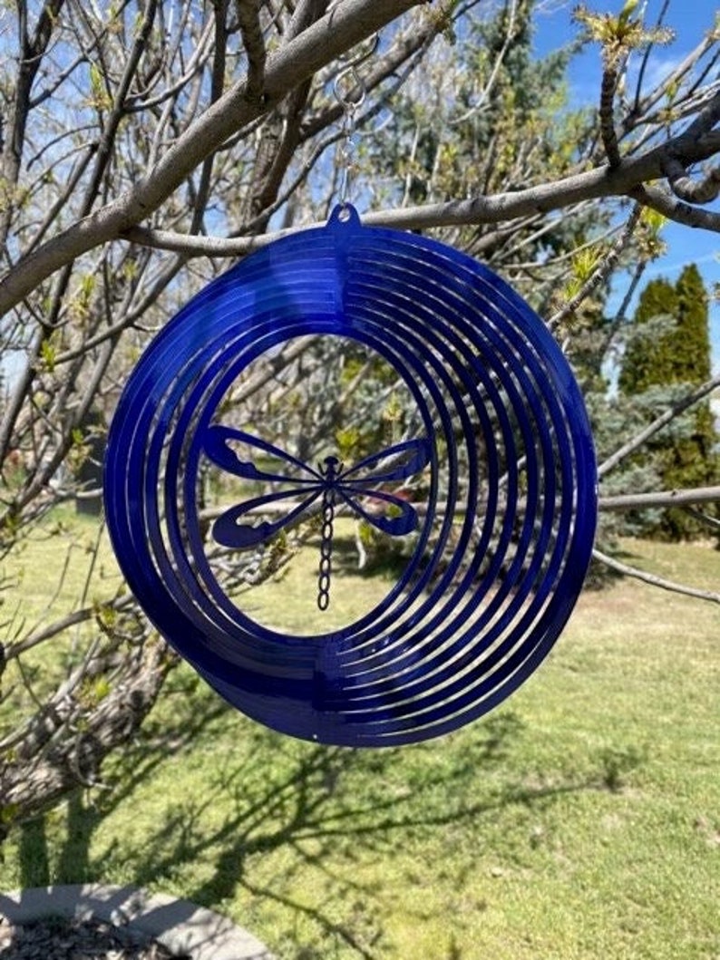 May include: A blue metal wind spinner with a dragonfly design. The spinner has multiple layers of metal that create a swirling effect when it spins in the wind.