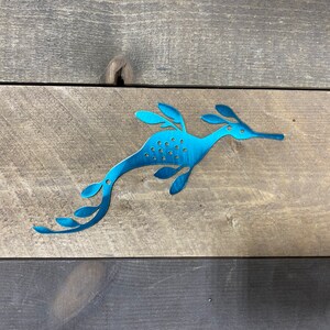 Op de afbeelding: Een tealblauwe metalen zeepaardje-uitsparing met een bladontwerp. Het zeepaardje kijkt naar rechts en heeft een lange snuit. De uitsparing bevindt zich op een houten achtergrond.