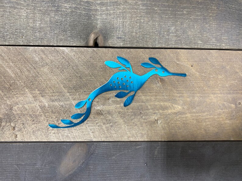 Op de afbeelding: Een tealblauwe metalen zeepaardje-uitsparing met een bladontwerp. Het zeepaardje kijkt naar rechts en heeft een lange snuit. De uitsparing bevindt zich op een houten achtergrond.