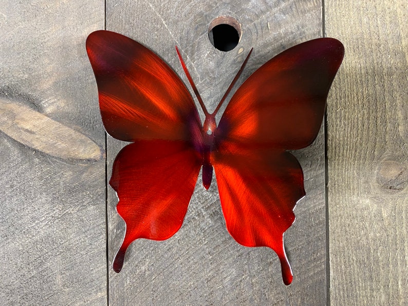 May include: A red metal butterfly wall decor with a dark red and black gradient on the wings. The butterfly is mounted on a wooden surface.