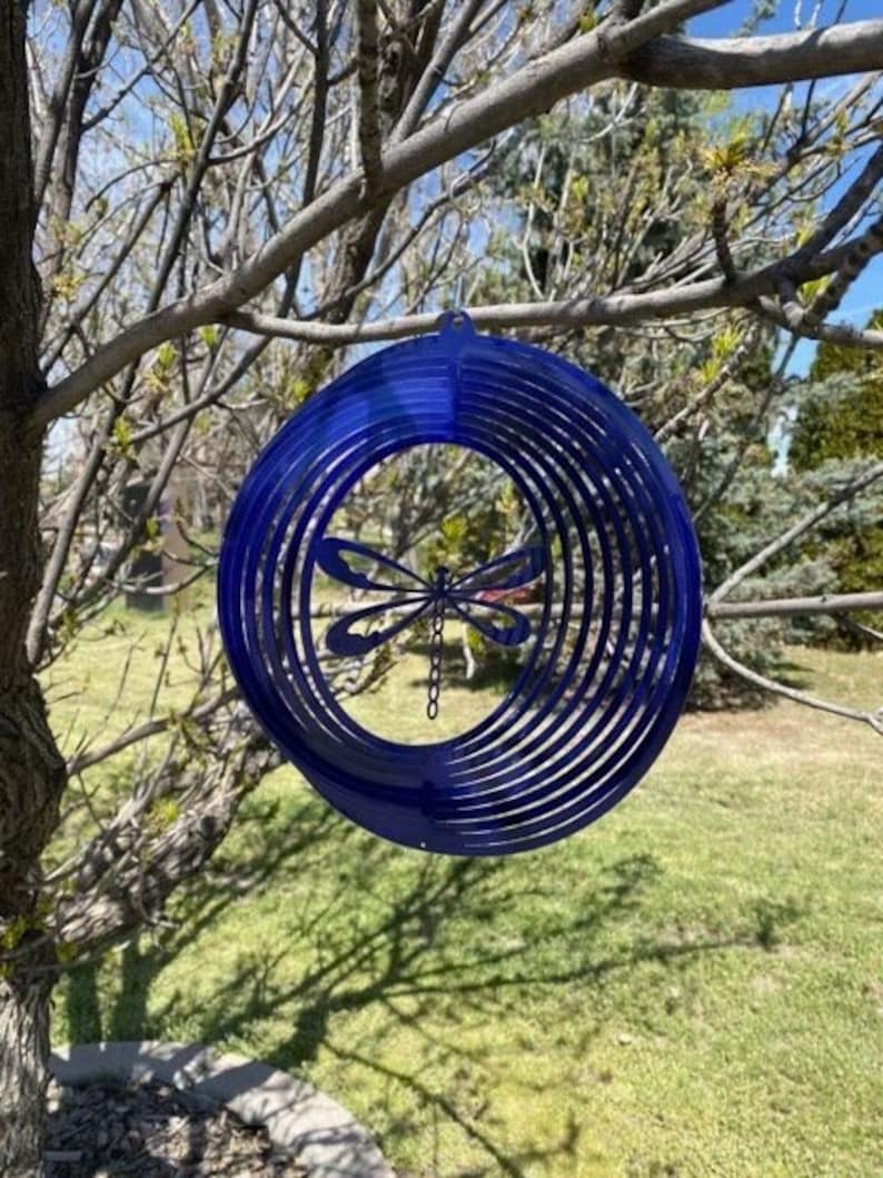 May include: A blue metal dragonfly wind spinner with multiple layers of circles. The spinner is hanging from a tree branch.