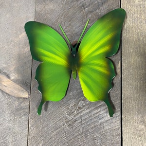 May include: A green metal butterfly wall decor with a yellow gradient. The butterfly is attached to a wooden surface.