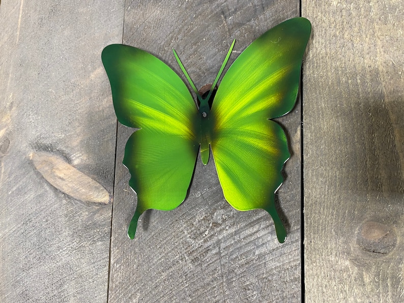 May include: A green metal butterfly wall decor with a yellow gradient. The butterfly is attached to a wooden surface.