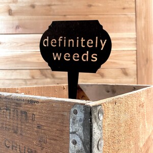 May include: Black metal sign with the text "definitely weeds" against a wooden backdrop.