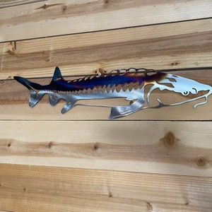 May include: A metal wall sculpture of a sturgeon fish with a blue, purple and orange iridescent finish. The fish is mounted on a light wood background.