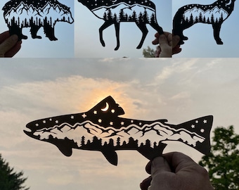 May include: A set of five black paper cutouts of animals and a fish, each with a mountain range and trees inside. The animals include a bear, an elk, a wolf, and a fish.