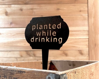 May include: Black metal garden marker with the text "planted while drinking" on a wooden background.