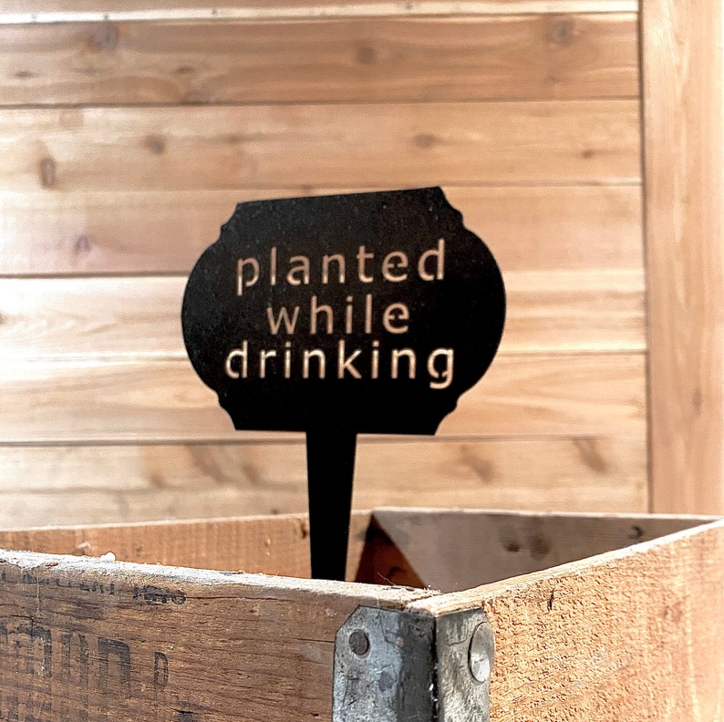 May include: Black metal garden marker with the text "planted while drinking" against a wooden backdrop.