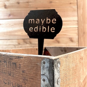 May include: A black metal sign with the text "maybe edible" on a wooden crate. The sign is attached to a metal stake.