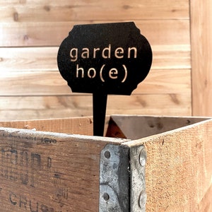 May include: Black metal garden marker with the text "garden ho(e)" in white lettering. The marker is inserted into a wooden crate.