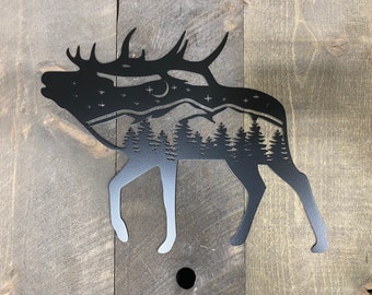 May include: Black metal silhouette of an elk with a mountain range and pine trees inside the silhouette. The elk is facing left and has large antlers.