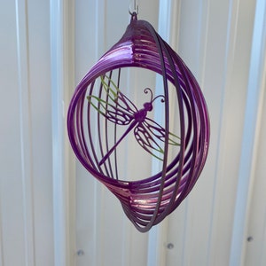 May include: Purple metal wind spinner featuring a dragonfly design. The spinner is crafted from multiple layers of metal, twisted and turned to create a swirling effect.