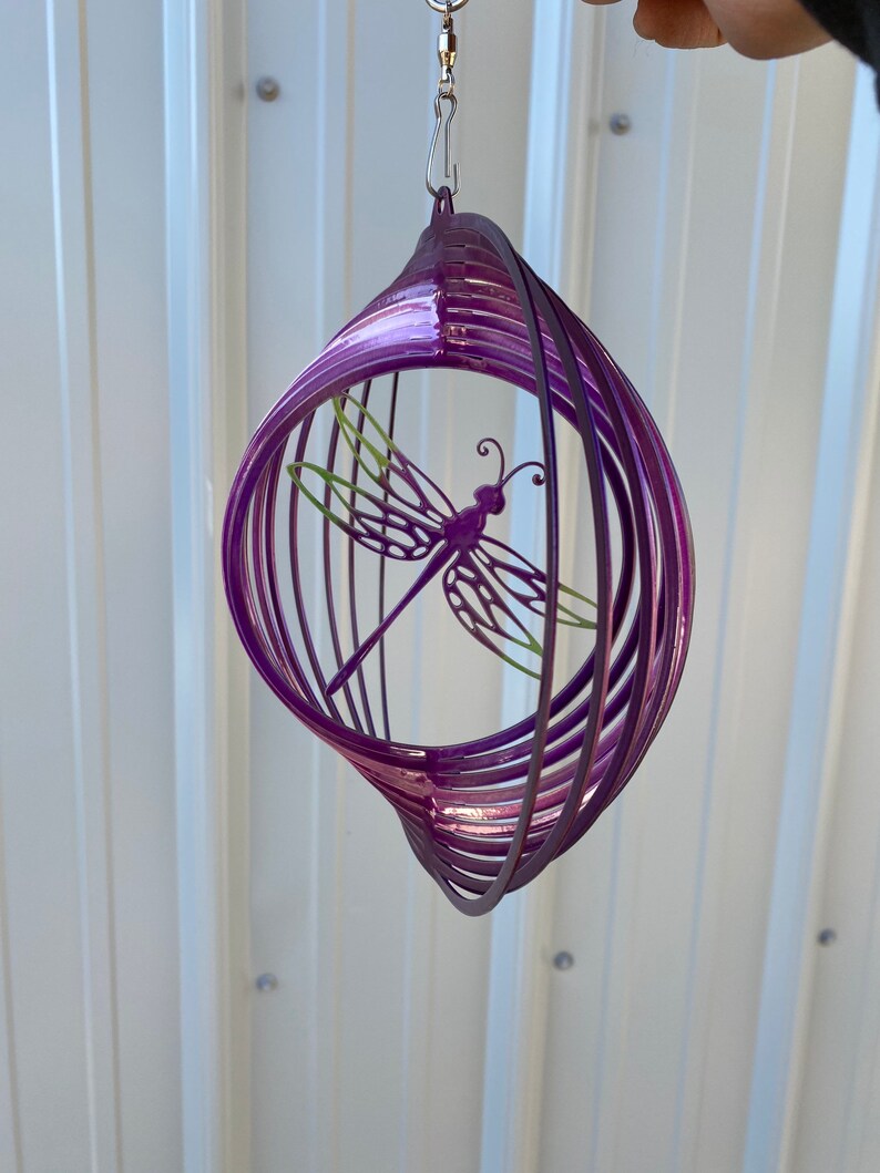May include: Purple metal wind spinner featuring a dragonfly design. The spinner is crafted from multiple layers of metal, twisted and turned to create a swirling effect.