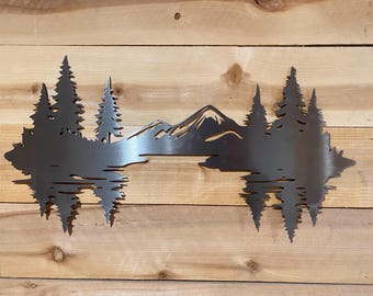 May include: Metal wall art depicting a mountain range and evergreen trees reflected in water. The artwork is cut from a single sheet of metal and mounted on a wooden background. The design features a detailed silhouette of the natural landscape.