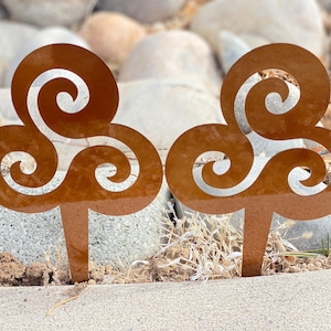 May include: Two rusty brown metal garden stakes with a swirling, clover-shaped design. The stakes are placed in the ground amongst rocks and dry grass.