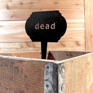 May include: A black metal sign with the word "dead" written in white letters, mounted on a wooden stake, placed in a wooden crate with metal corner brackets.