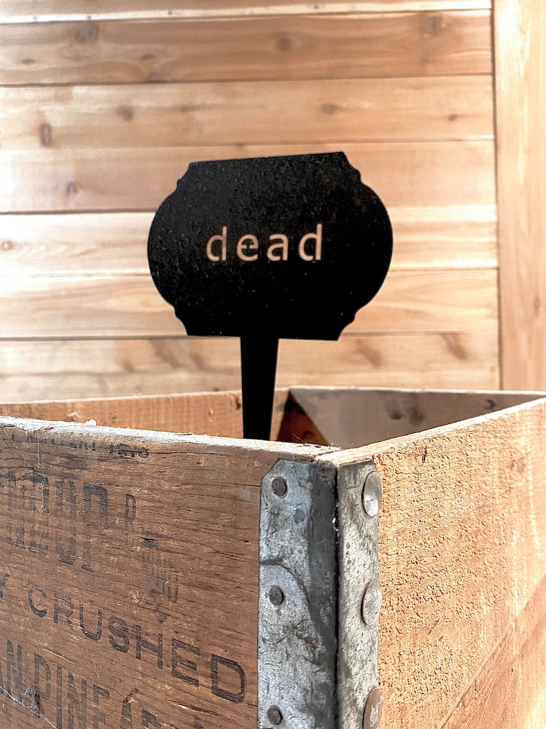 May include: A black metal sign with the word "dead" written in white letters, mounted on a wooden stake, placed in a wooden crate with metal corner brackets.
