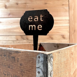 May include: A black metal sign with the words "eat me" in gold lettering. The sign is attached to a wooden stick and is sitting in a wooden crate.