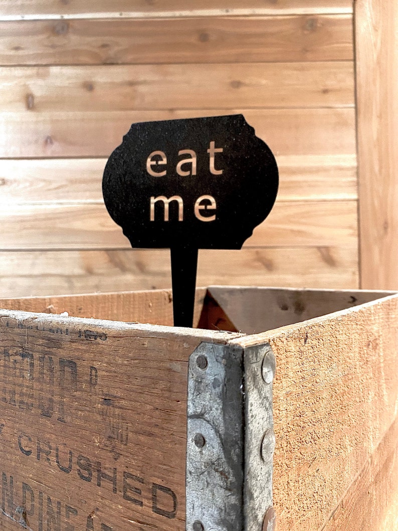 May include: A black metal sign with the words "eat me" in gold lettering. The sign is attached to a wooden stick and is sitting in a wooden crate.