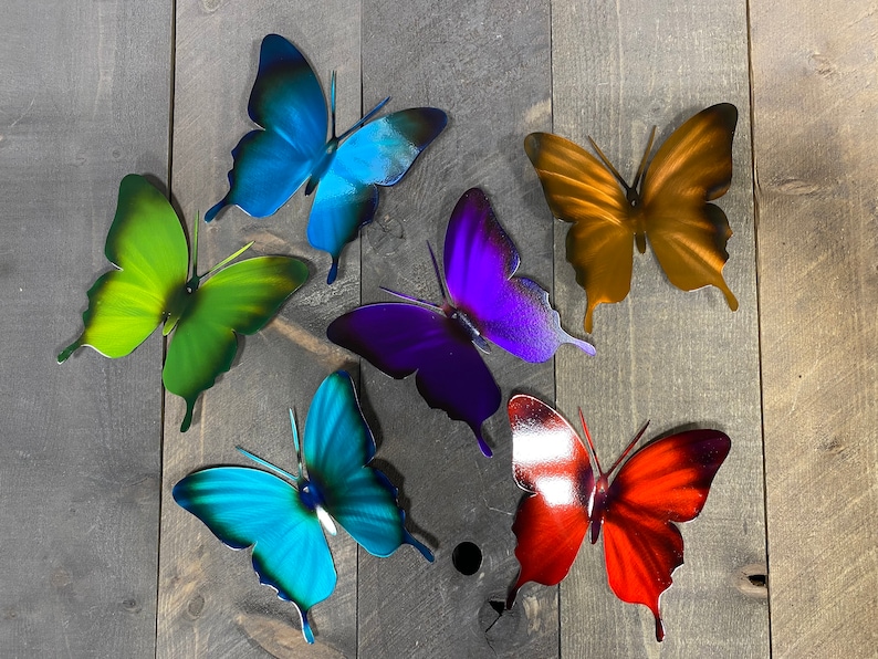 May include: Six metal butterflies in various colors, including green, blue, purple, orange, red, and turquoise, are displayed on a wood background.