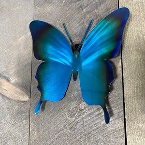 May include: A blue metal butterfly wall decor with a gradient of dark blue to light blue. The butterfly is attached to a wooden surface.