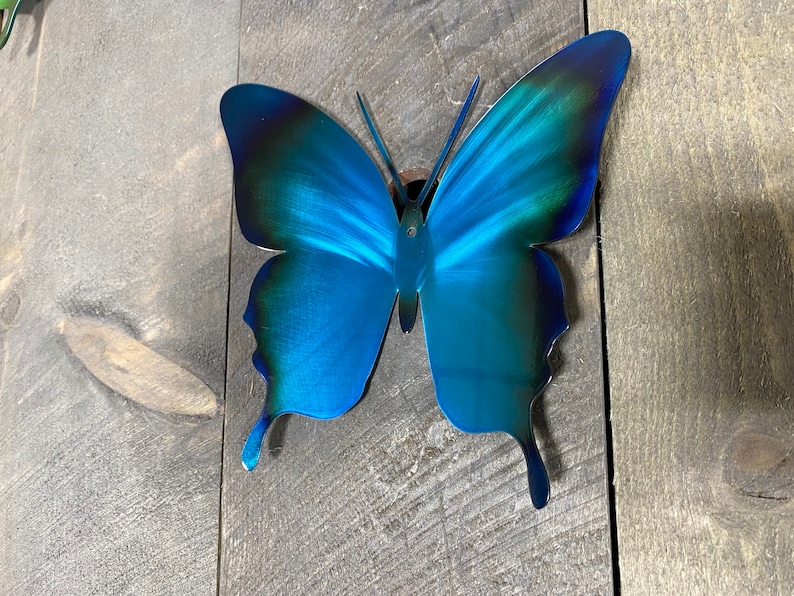 May include: A blue metal butterfly wall decor with a gradient of dark blue to light blue. The butterfly is attached to a wooden surface.