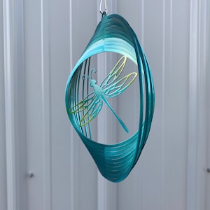 May include: A teal blue metal wind spinner featuring a dragonfly design. The dragonfly is cut out of the metal and has a green and yellow gradient.