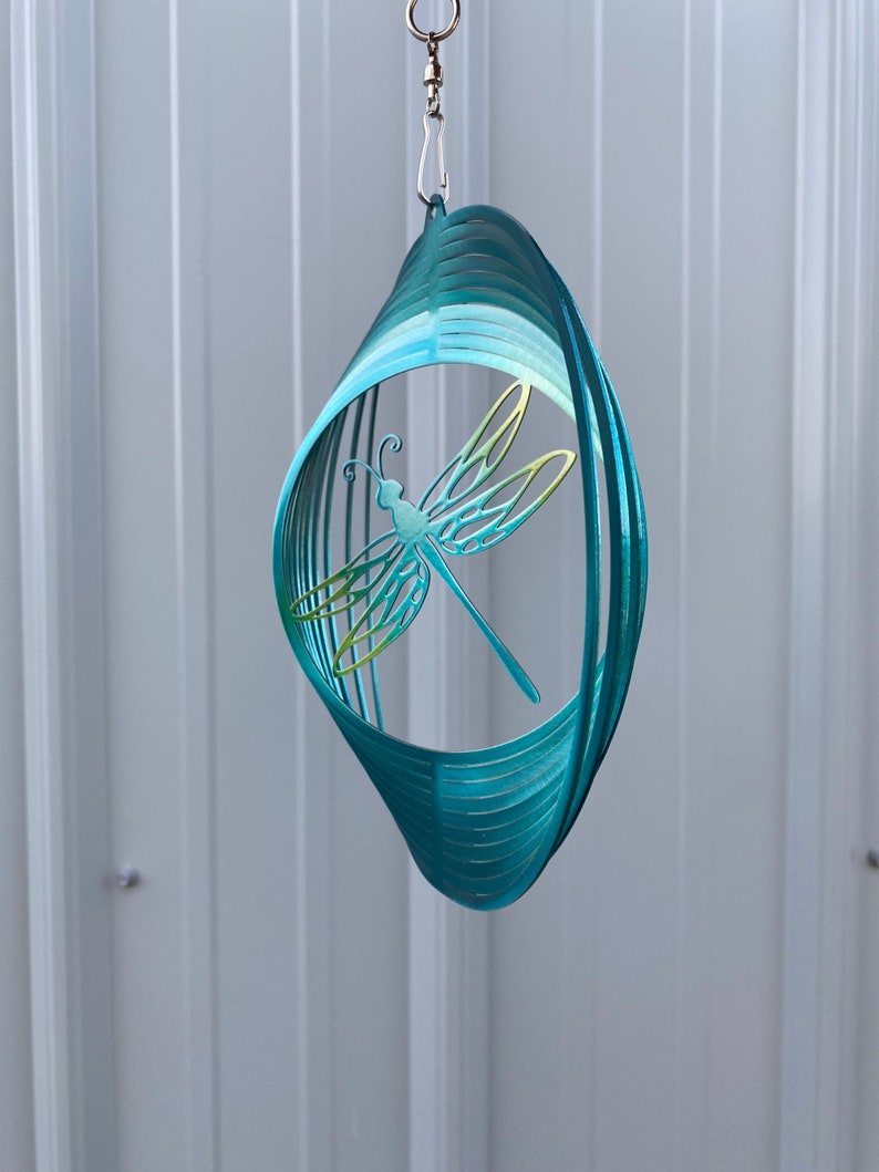 May include: A teal blue metal wind spinner featuring a dragonfly design. The dragonfly is cut out of the metal and has a green and yellow gradient.