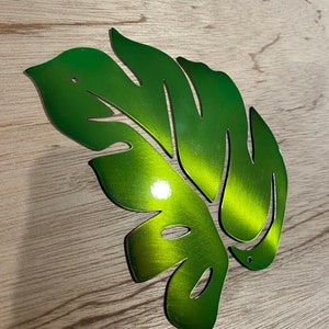 Puede incluir: Una hoja de metal verde con un dise&ntilde;o recortado, que se asemeja a una hoja de monstera. La hoja tiene un acabado met&aacute;lico brillante.
