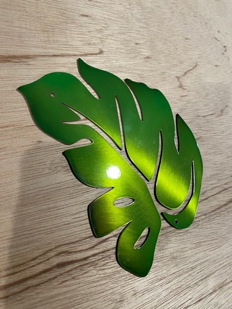 Puede incluir: Una hoja de metal verde con un dise&ntilde;o recortado, que se asemeja a una hoja de monstera. La hoja tiene un acabado met&aacute;lico brillante.