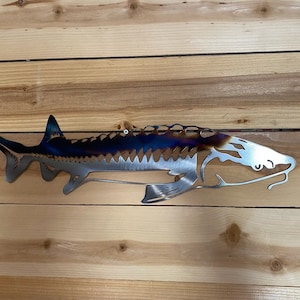 May include: A metal wall sculpture of a sturgeon fish with a blue and brown patina finish. The fish is depicted in a side profile with its mouth open and its fins spread out. The sculpture is made of metal and has a rustic, industrial look.