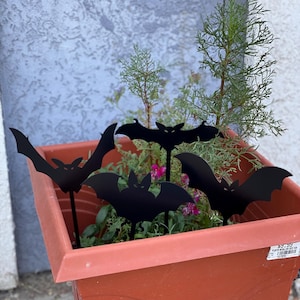 May include: Four black metal bat silhouettes are placed in a terracotta pot filled with green plants. The bats are on stakes and are facing different directions. The pot has a price tag attached to it that reads "$5.99".