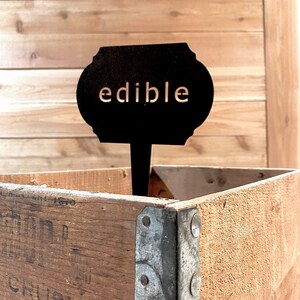 May include: Black metal sign with rounded edges and the word "edible" in white lettering. The sign is attached to a wooden stick and is sitting in a wooden crate.