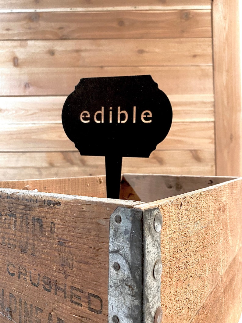 May include: Black metal sign with rounded edges and the word "edible" in white lettering. The sign is attached to a wooden stick and is sitting in a wooden crate.