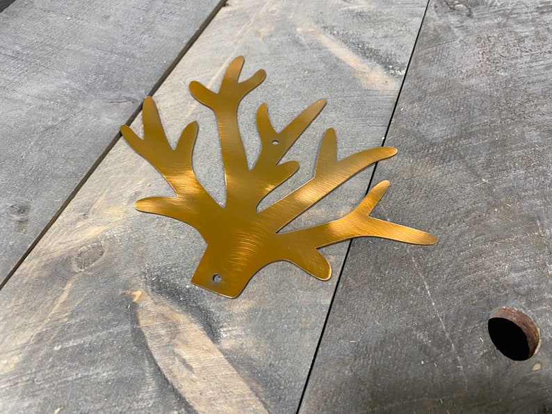 Puede incluir: Un recorte de metal dorado de una rama de &aacute;rbol estilizada con m&uacute;ltiples ramas. El recorte est&aacute; sobre una superficie de madera gris.