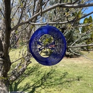 May include: A blue metal dragonfly wind spinner with a circular design. The spinner is hanging from a tree branch.