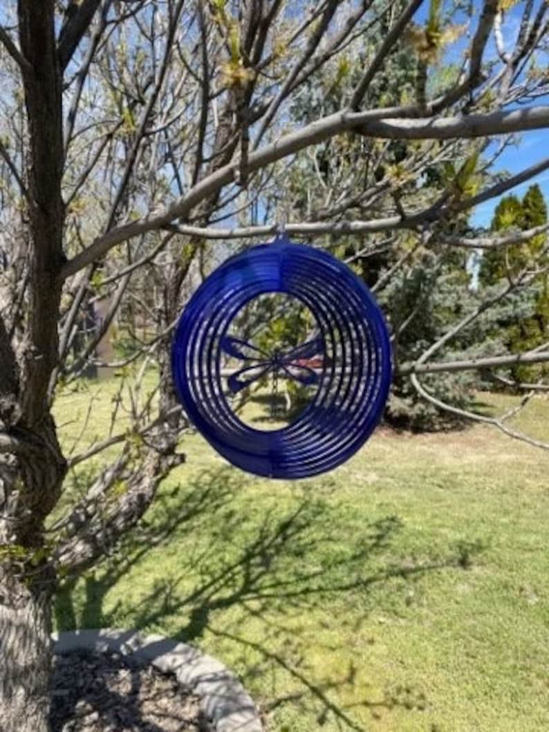 May include: A blue metal dragonfly wind spinner with a circular design. The spinner is hanging from a tree branch.
