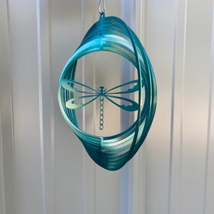 May include: Teal metal dragonfly wind spinner with a circular design. The spinner is suspended from a silver chain.