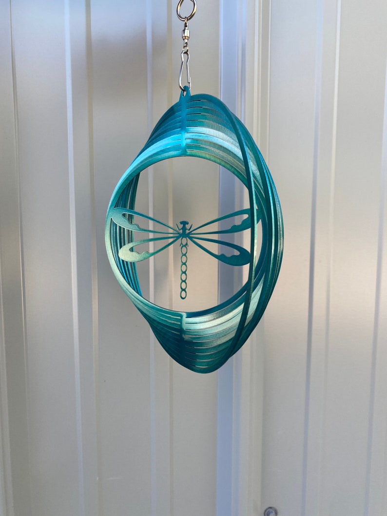 May include: Teal metal dragonfly wind spinner with a circular design. The spinner is suspended from a silver chain.