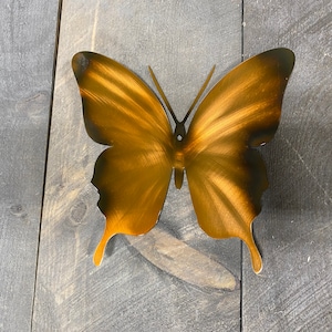 May include: A gold metal butterfly wall decor with a brushed finish. The butterfly is mounted on a gray wooden surface.