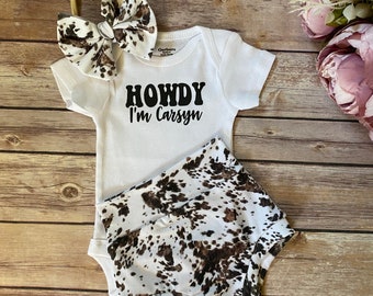 May include: A white onesie with the text "Howdy I'm Carsyn" in black. The onesie is paired with brown and white cow print shorts and a brown and white cow print headband.