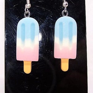May include: A pair of earrings shaped like popsicles. The popsicles are blue, white, and pink with a yellow stick.
