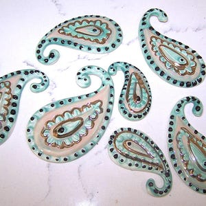 May include: Seven ceramic paisley-shaped ornaments. Each ornament features a light blue border, a pink and gold interior, and black dot accents. The ornaments have a glossy finish and a curved, swirled top.